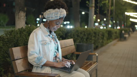 Mujer-Afroamericana-Sentada-En-El-Parque-Por-La-Noche-Y-Usando-Una-Laptop